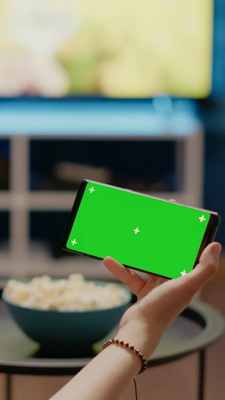 Dłoń trzyma smartfon z pustym zielonym ekranem w układzie poziomym na tle telewizora