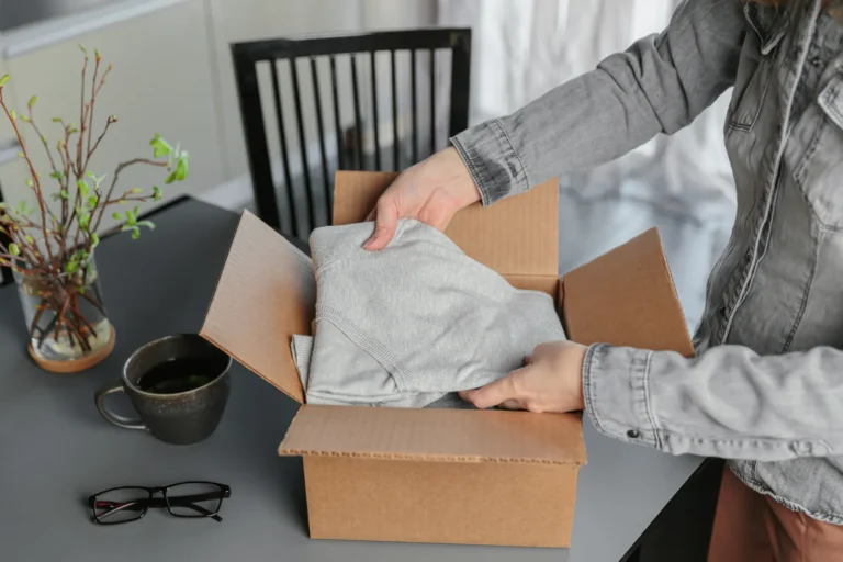 Kobieta wyjmuje sweter z kartonowej paczki przy biurku — zwrot zamówienia ze sklepu internetowego