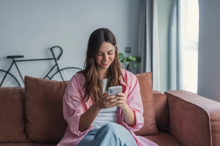 Uśmiechnięta kobieta robi zakupy w aplikacji na smartfonie, siedząc na kanapie