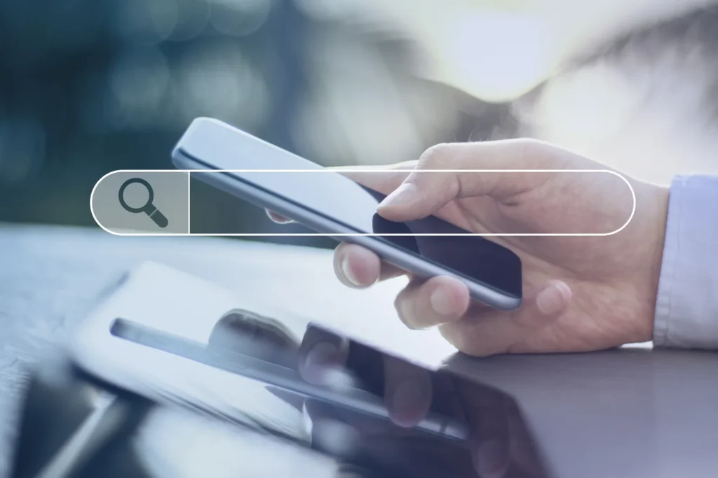 Dłoń trzyma smartfon, nad nim grafika paska wyszukiwania z lupą